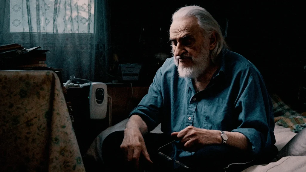 old man in a blue shirt sitting lost in thought on his bed with his glasses in his hand at the end of life